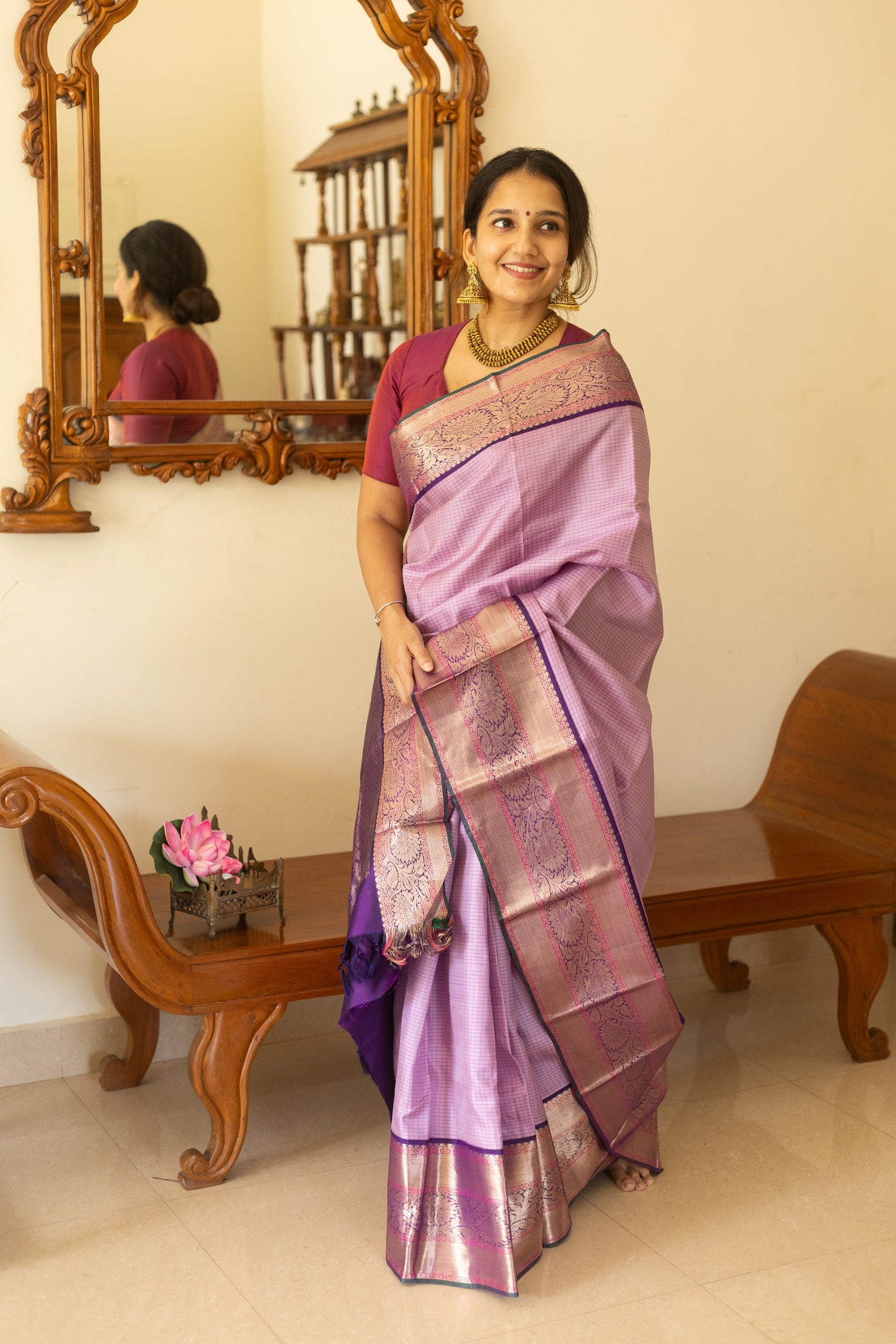 Woman in a purple saree standing in a room with wooden furniture and a mirror.