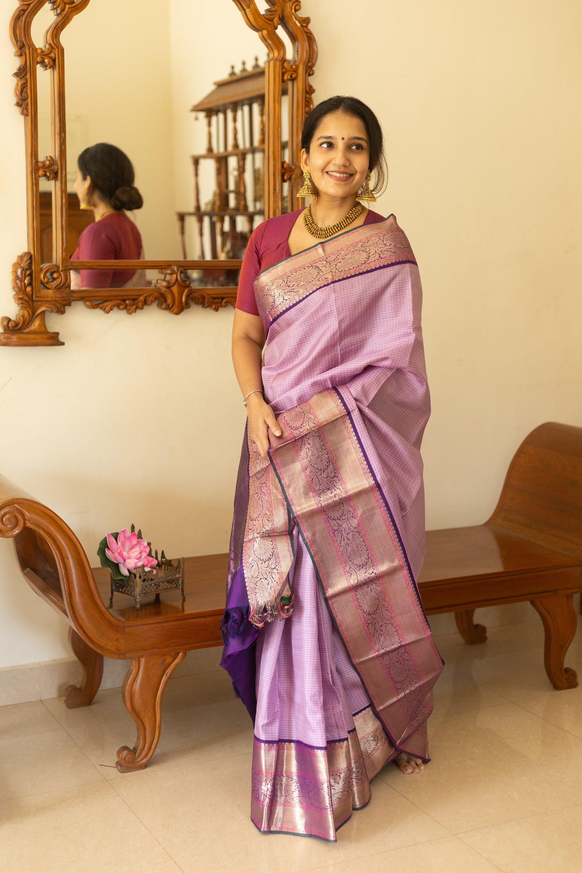 Woman in a purple saree standing in a room with wooden furniture and a mirror.